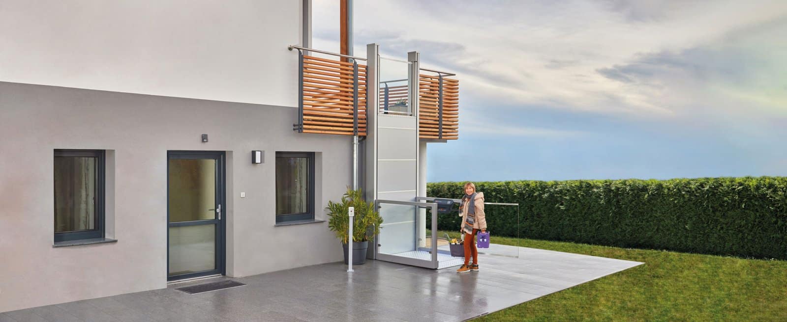 Ascendor_UP 25 Modern house exterior with a wooden slatted balcony and glass railing; a woman stands on the patio with shopping bags.