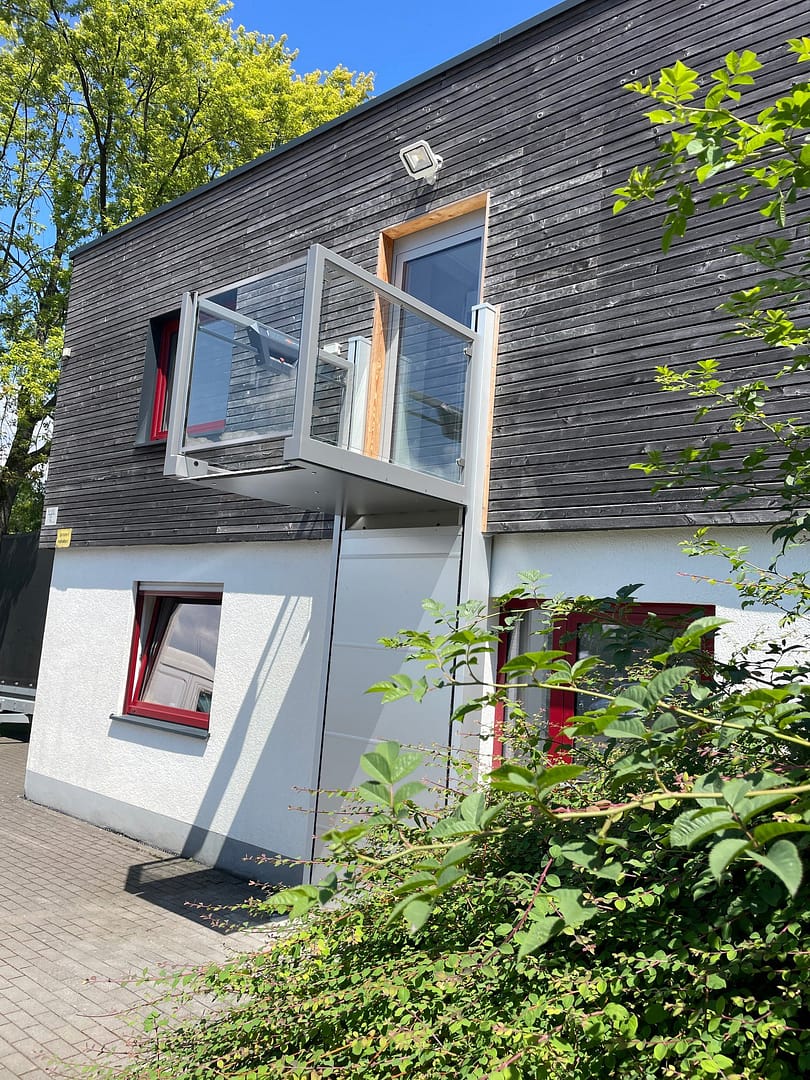 Ascendor_UP 26 Modern two-story building with dark wooden siding, glass balcony railing, and red-framed windows under a blue sky, with greenery in foreground