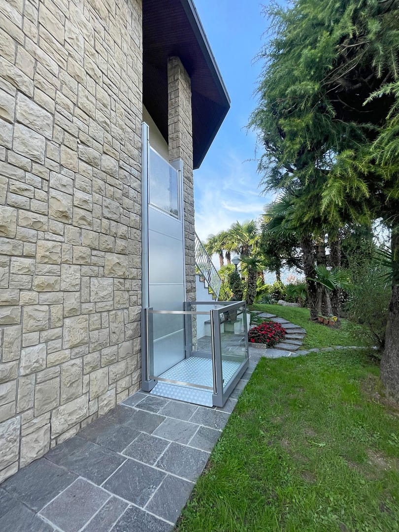Ascendor_UP 6 Stone-clad house exterior with a glass door and metal railing, set on a stone tile path beside a green lawn and tall trees.