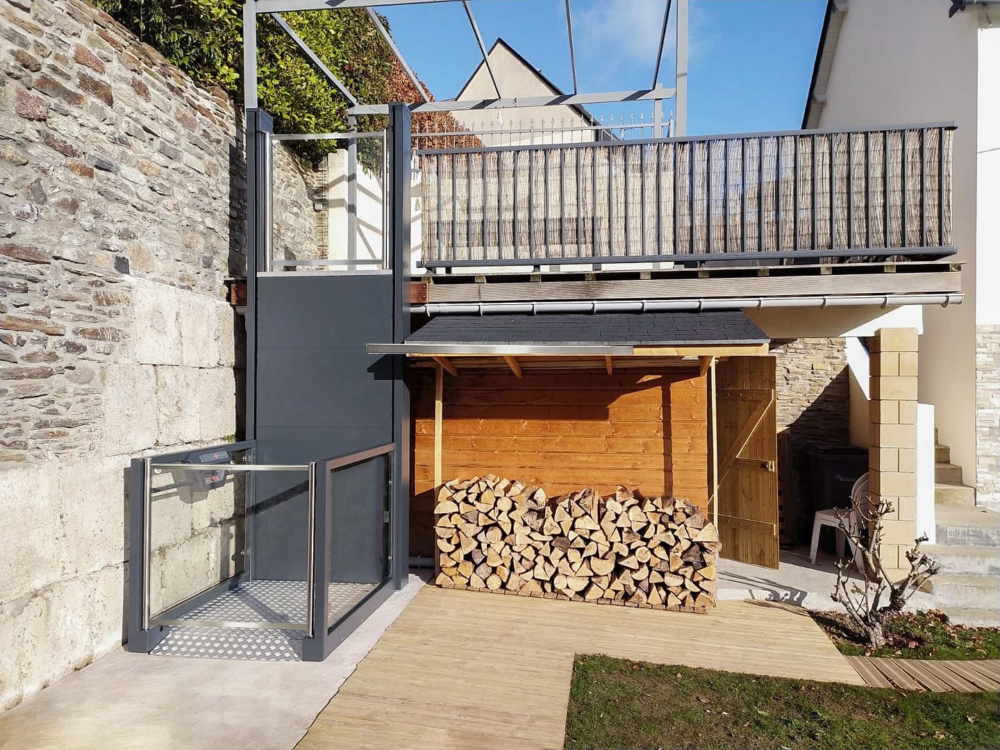 Ascendor_UP 21 ‣ Serenika Wood storage area under a balcony with stacked firewood and a wooden shed, metal railing, and stone walls surrounding the yard.