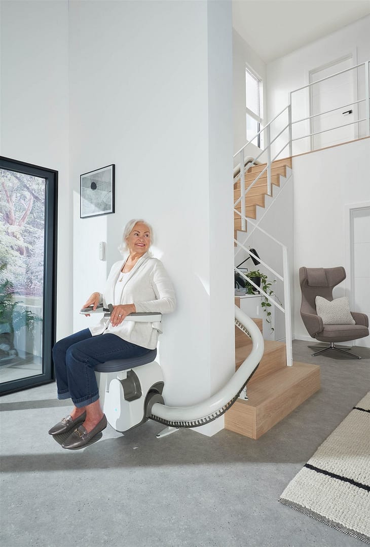 Femme assise sur le monte-escalier Flow X au pied d'un escalier tournant, rail courbe blanc
